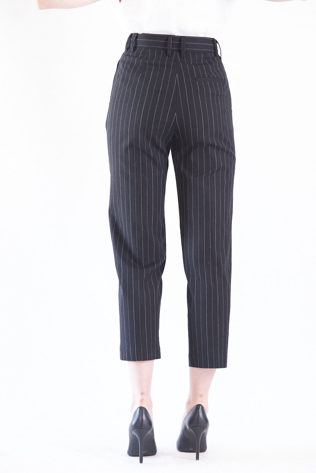 A woman exudes confidence in Le Réussi®s Power Woman- Black/White Stripes Pants paired with a white blouse, standing stylishly with one hand in her pocket against a white background.