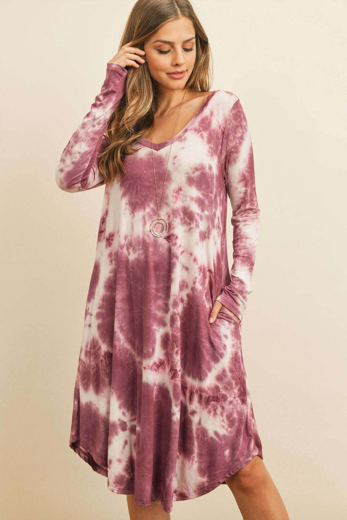 A woman in a purple Tie Dye V-Neck Rounded Hem Midi Dress by Riah Fashion, touching her hair, stands against a beige background.