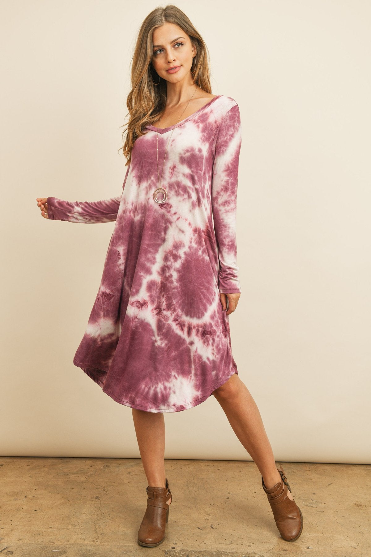 A woman in a purple Tie Dye V-Neck Rounded Hem Midi Dress by Riah Fashion, touching her hair, stands against a beige background.