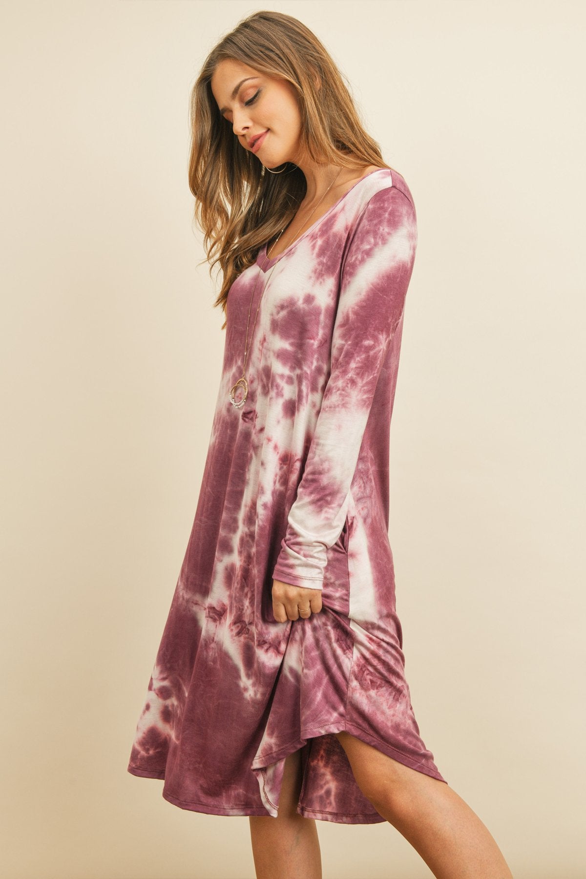 A woman in a purple Tie Dye V-Neck Rounded Hem Midi Dress by Riah Fashion, touching her hair, stands against a beige background.