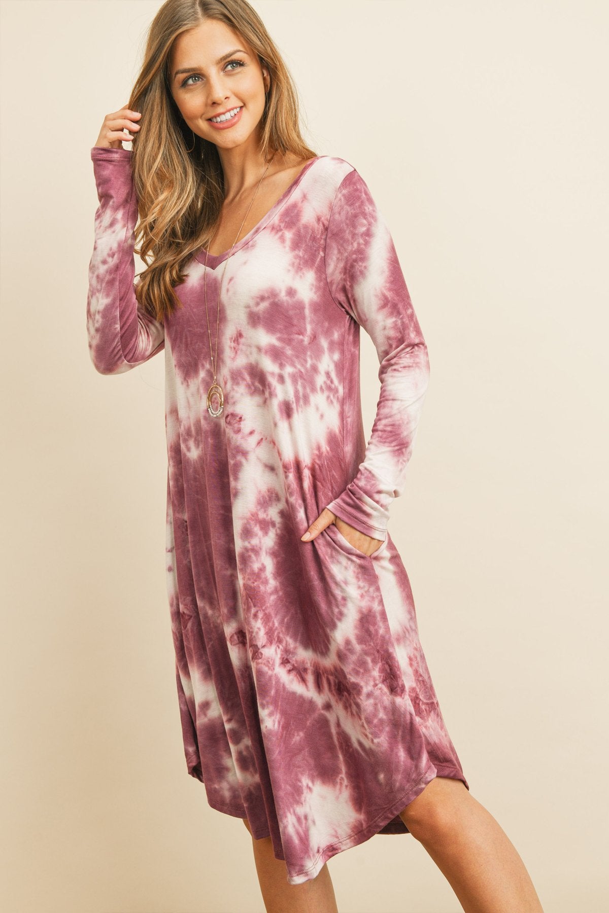 A woman in a purple Tie Dye V-Neck Rounded Hem Midi Dress by Riah Fashion, touching her hair, stands against a beige background.