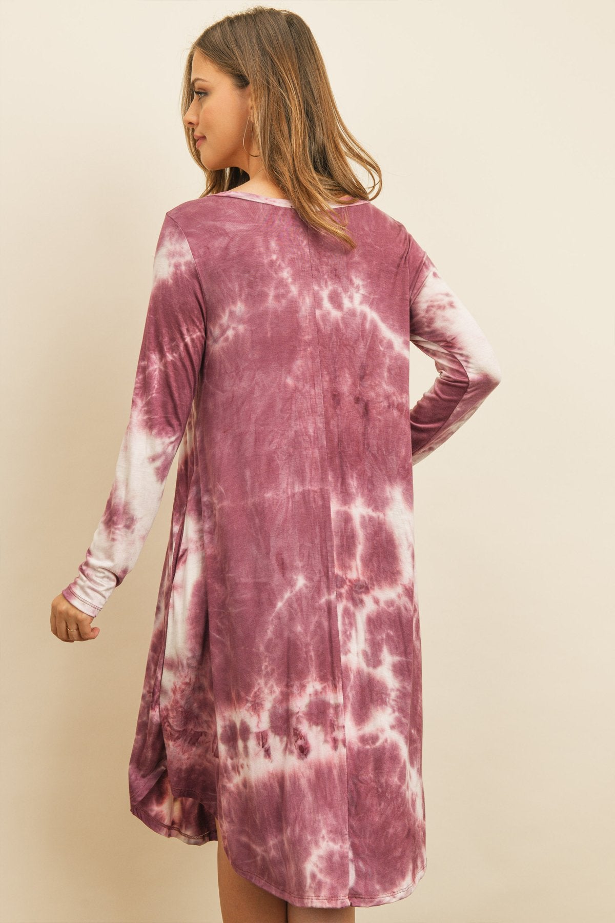 A woman in a purple Tie Dye V-Neck Rounded Hem Midi Dress by Riah Fashion, touching her hair, stands against a beige background.