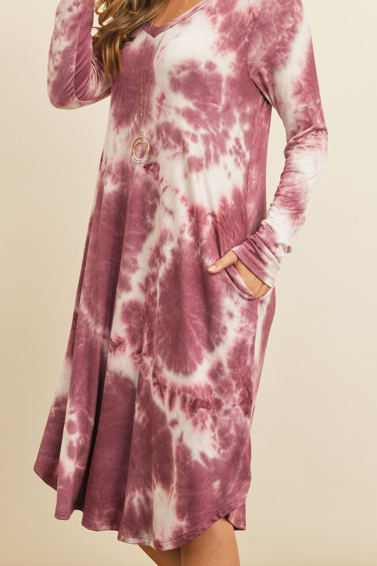 A woman in a purple Tie Dye V-Neck Rounded Hem Midi Dress by Riah Fashion, touching her hair, stands against a beige background.