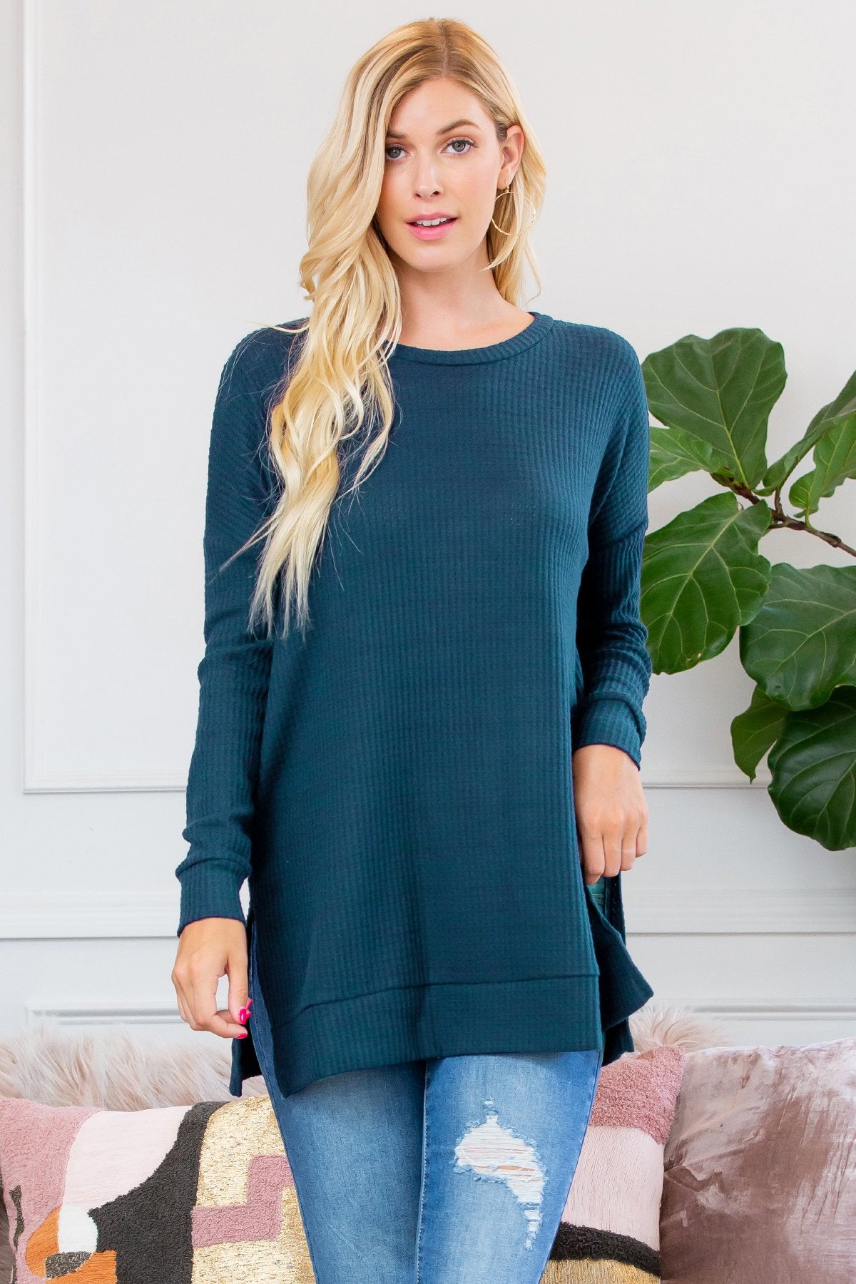 A woman stands on a wooden porch wearing Riah Fashions Brushed Thermal Waffle Sweater and jeans, accessorized with a necklace. Greenery and a wooden fence are visible in the background.