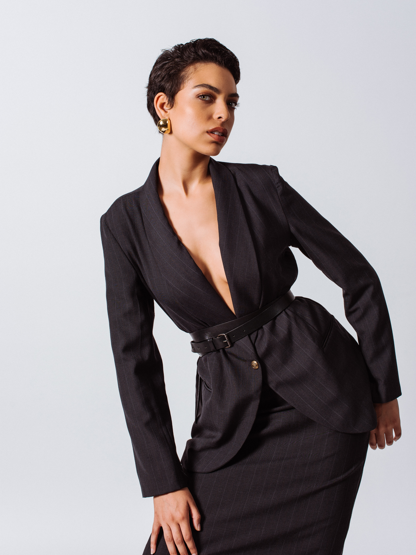 A person with short blond hair poses confidently in a beige top, skirt, and the Tailor Wool Dark Grey Blazer by Le Réussi® against a white background.