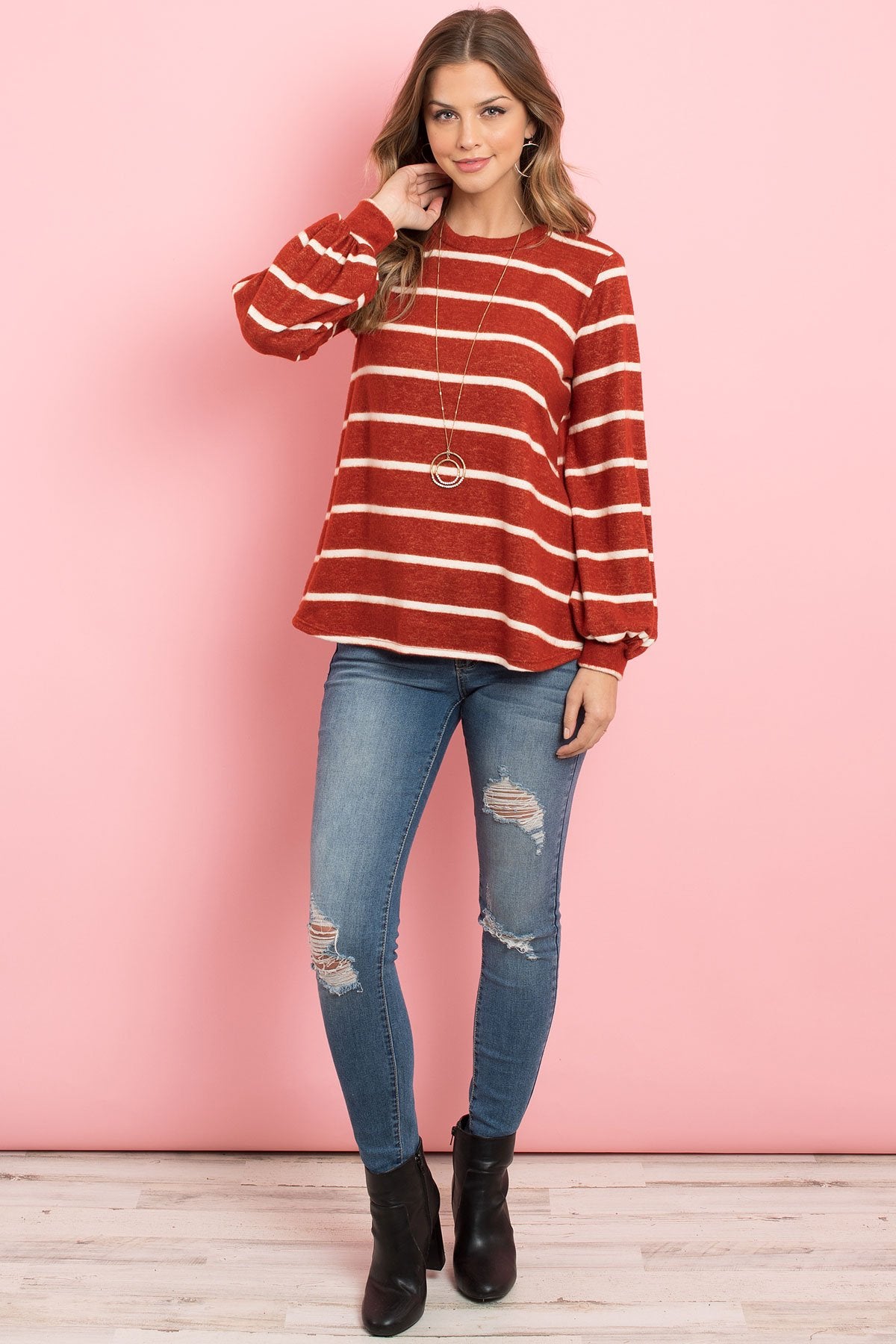A woman in a Riah Fashion Striped Long Sleeved Round Neck Swing Top and jeans stands against a pink backdrop, wearing a long necklace and touching her hair.