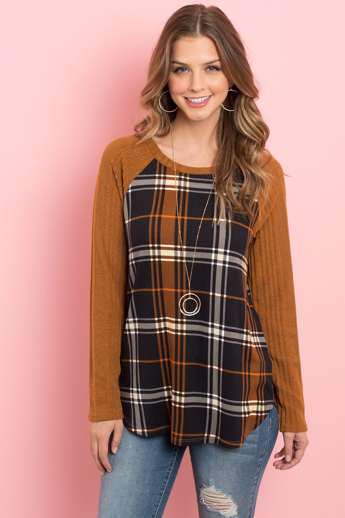 A person wearing a Riah Fashion Long Sleeve Knit Detail Contrast Plaid Top and jeans stands against a pink background, smiling.