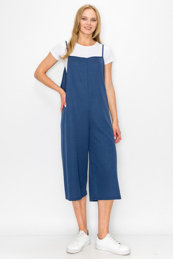 A person wearing a Stylespect Cropped Bottom Wide Leg Oversized Jumpsuit in blue over a white T-shirt and white sneakers, standing and smiling against a plain white background.