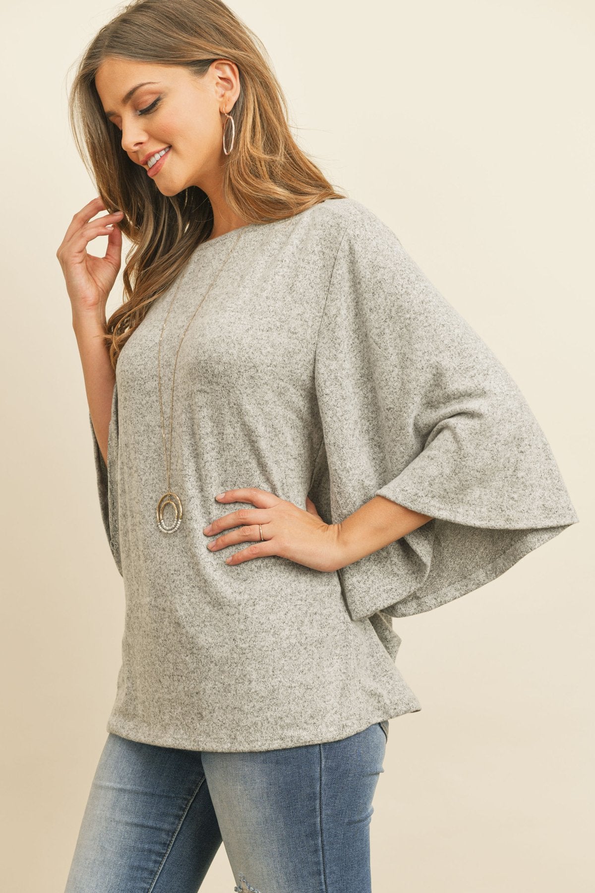 A person in a Riah Fashion Boat Neck Bell Sleeve Solid Hacci Brushed Top and ripped jeans stands against a neutral background, accessorized with hoop earrings and a long necklace.