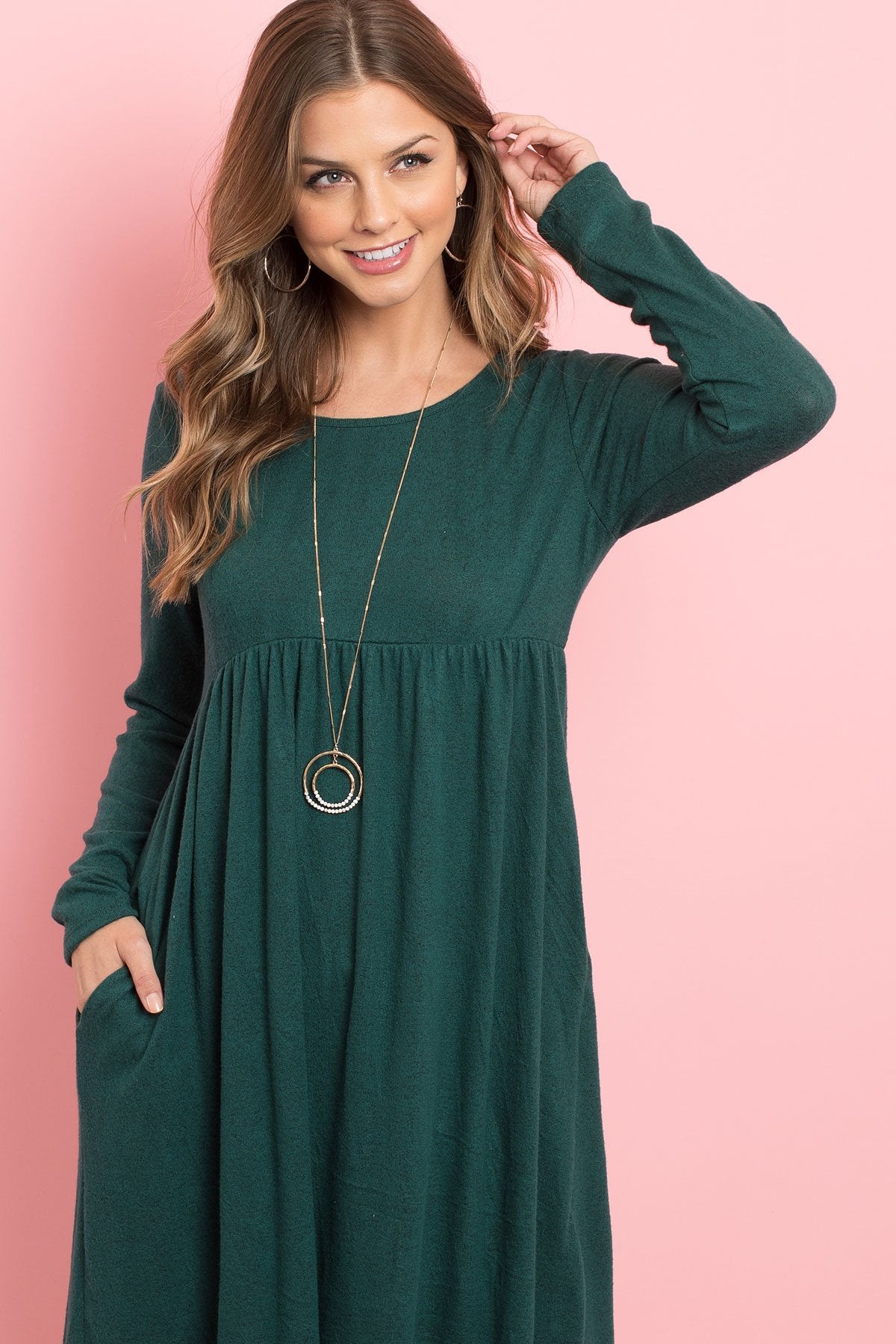 A woman in a Riah Fashion Boat Neck Brushed Hacci Round Hem Dress in Dark Ruby stands before a pink backdrop, adorned with a long necklace featuring circular pendants.