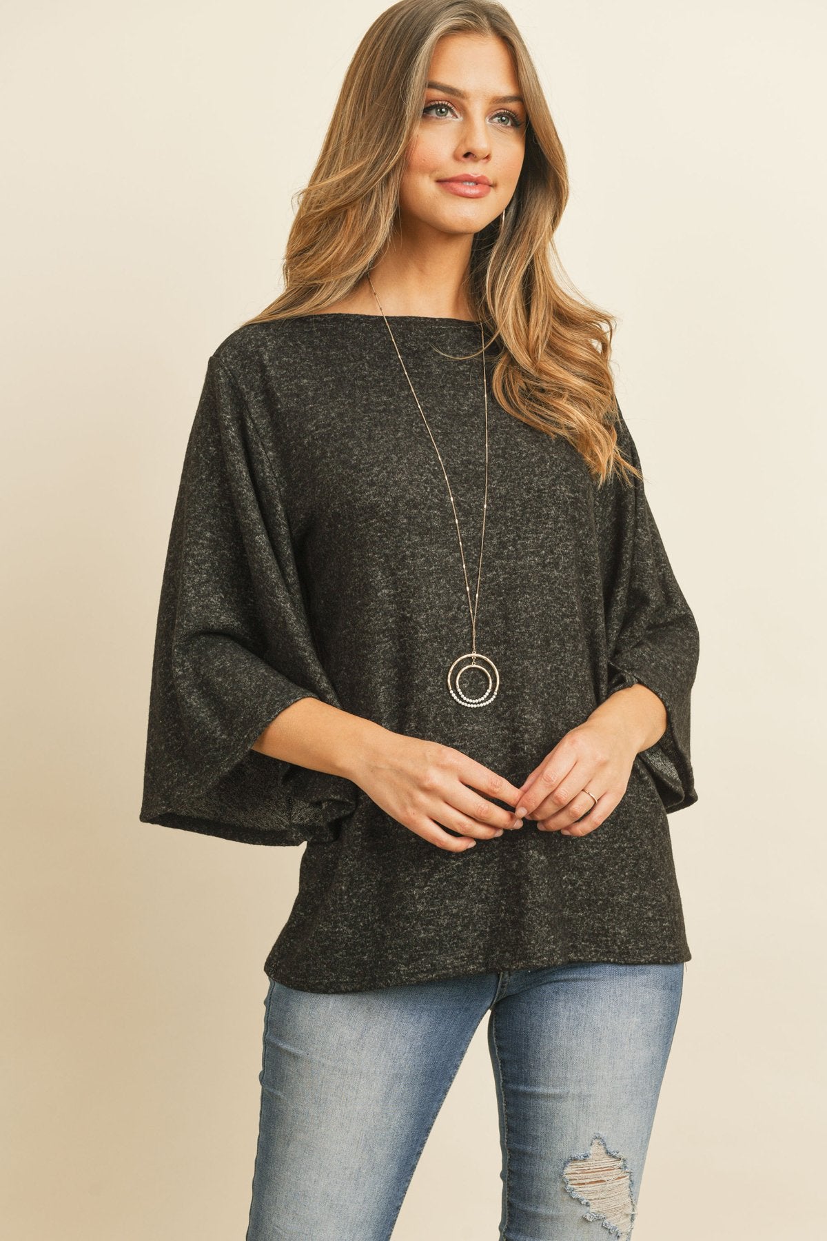 A person in a Riah Fashion Boat Neck Bell Sleeve Solid Hacci Brushed Top and ripped jeans stands against a neutral background, accessorized with hoop earrings and a long necklace.