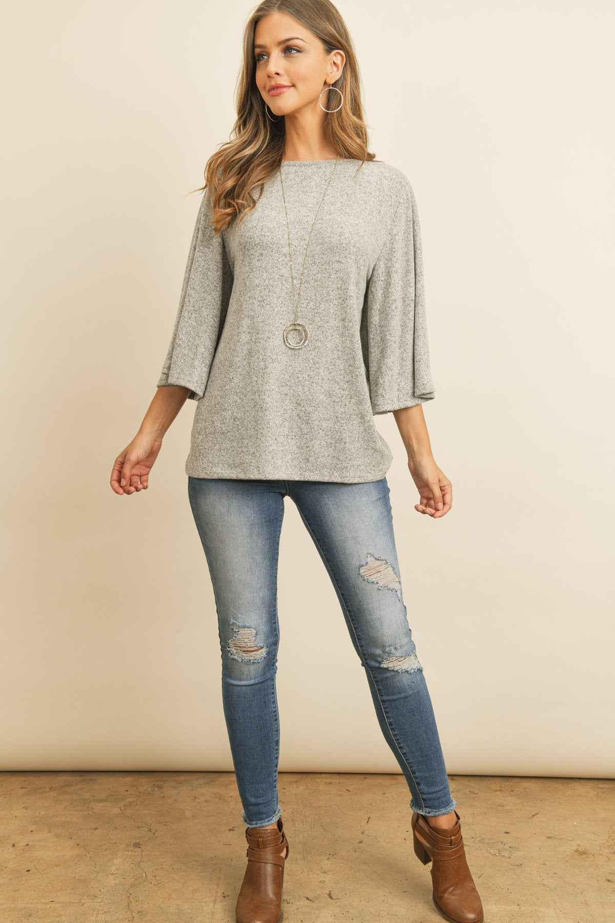 A person in a Riah Fashion Boat Neck Bell Sleeve Solid Hacci Brushed Top and ripped jeans stands against a neutral background, accessorized with hoop earrings and a long necklace.