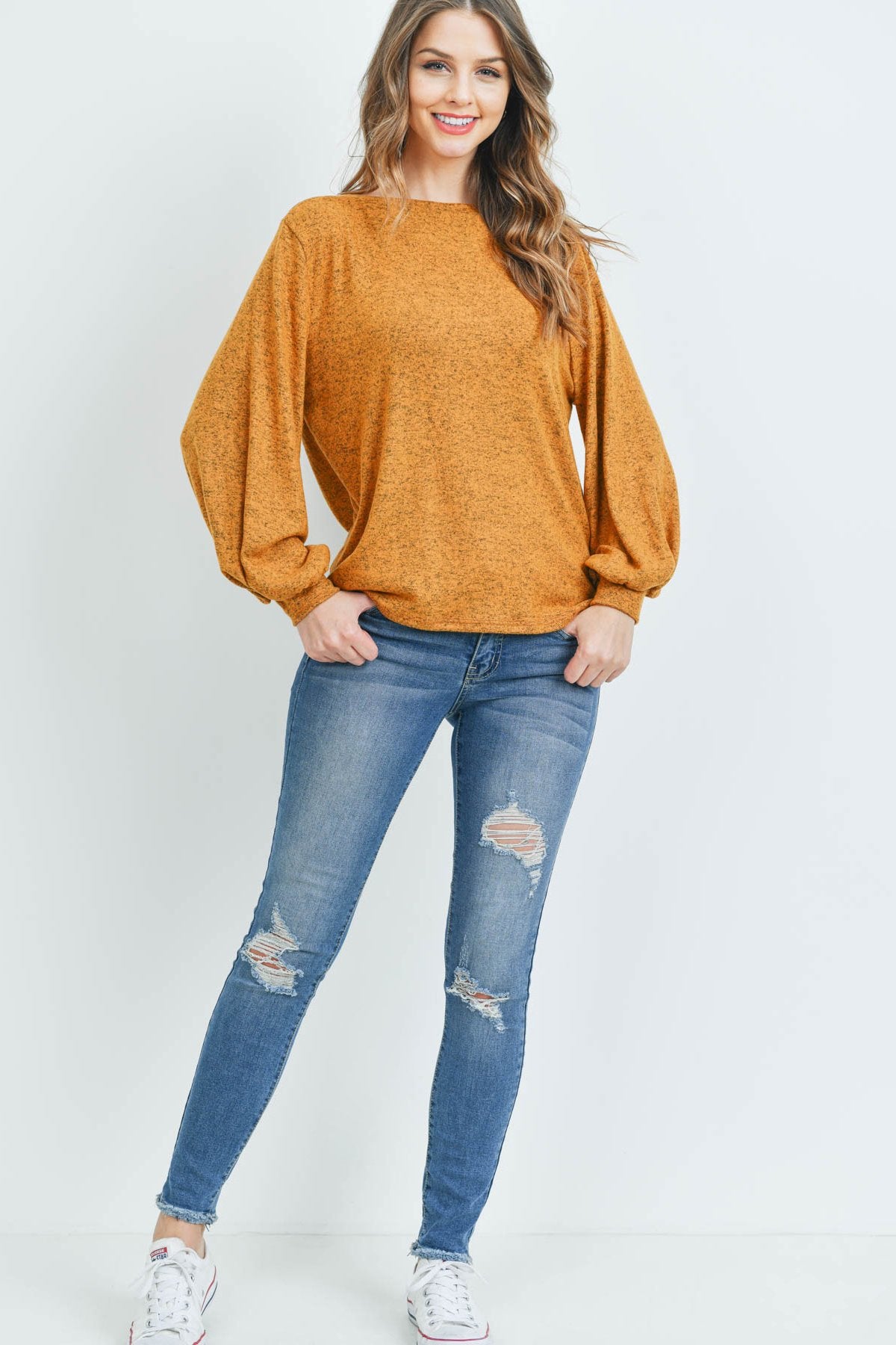A woman models a Riah Fashion Puff Sleeved Boat Neck Two Toned Brushed Hacci Top with ripped jeans and brown boots on a concrete floor against a plain backdrop, accessorized with a long necklace.