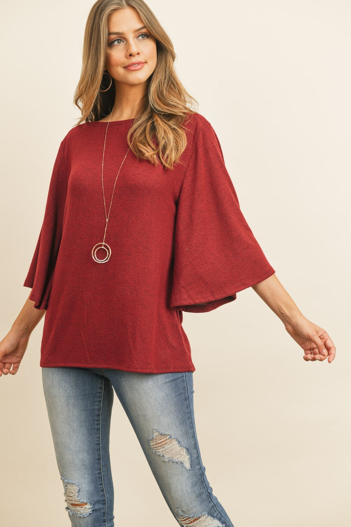 A person in a Riah Fashion Boat Neck Bell Sleeve Solid Hacci Brushed Top and ripped jeans stands against a neutral background, accessorized with hoop earrings and a long necklace.