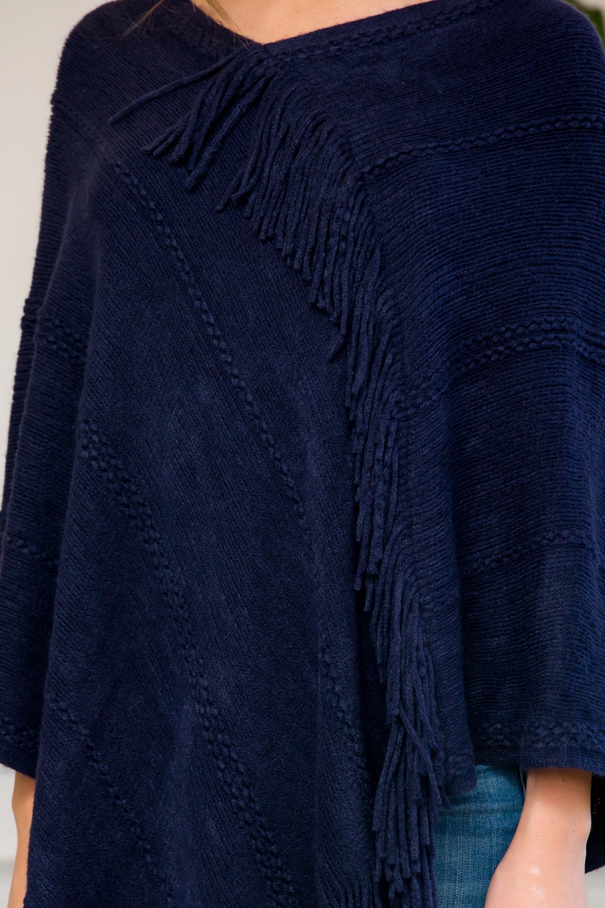 A person stands wearing a Riah Fashion Asymmetric Fringe Poncho in one color, paired with jeans and brown boots, smiling while holding the poncho. In the background are a plant and a sofa with cushions.