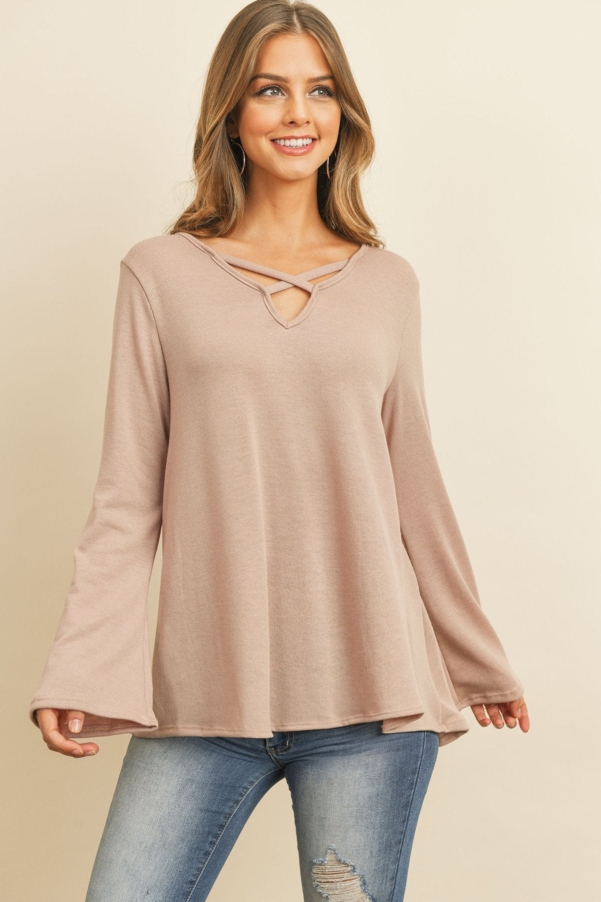A person wearing a Riah Fashion Criss Cross Neck Long Sleeve Hacci Top in Dusty Blush and blue jeans stands against a neutral background, smiling.