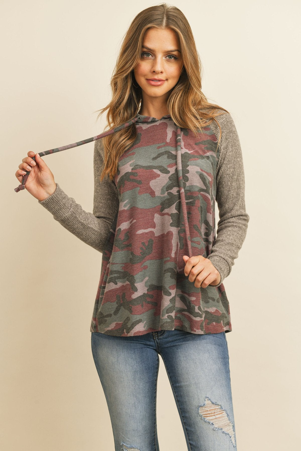 A person wearing the Self Tie Rib Hacci Brushed Sleeve Camouflage Hoodie by Riah Fashion with distressed jeans, holding the hoodie strings, stands against a beige background.