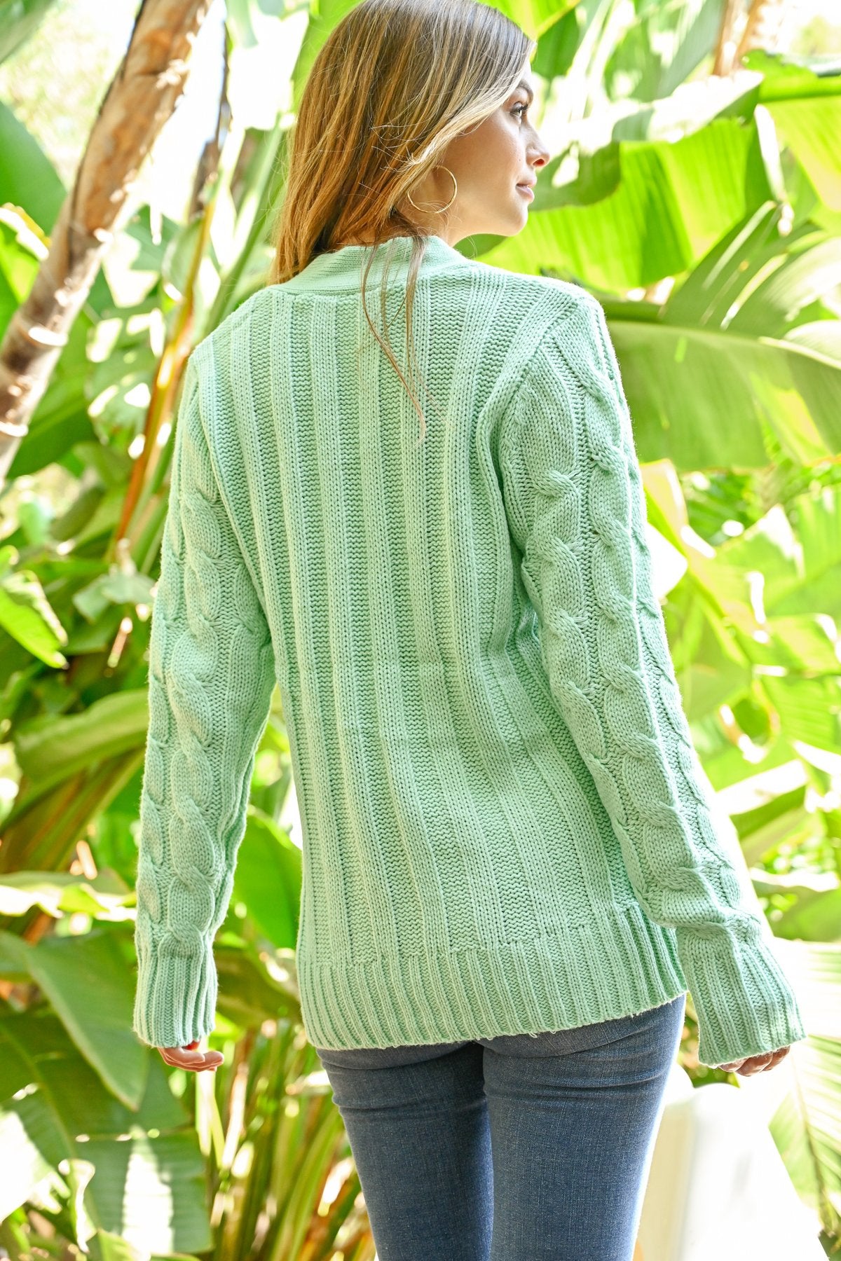 A person wearing an ash copper cable sweater with pockets from Riah Fashion stands in front of tall green plants and a wooden fence.