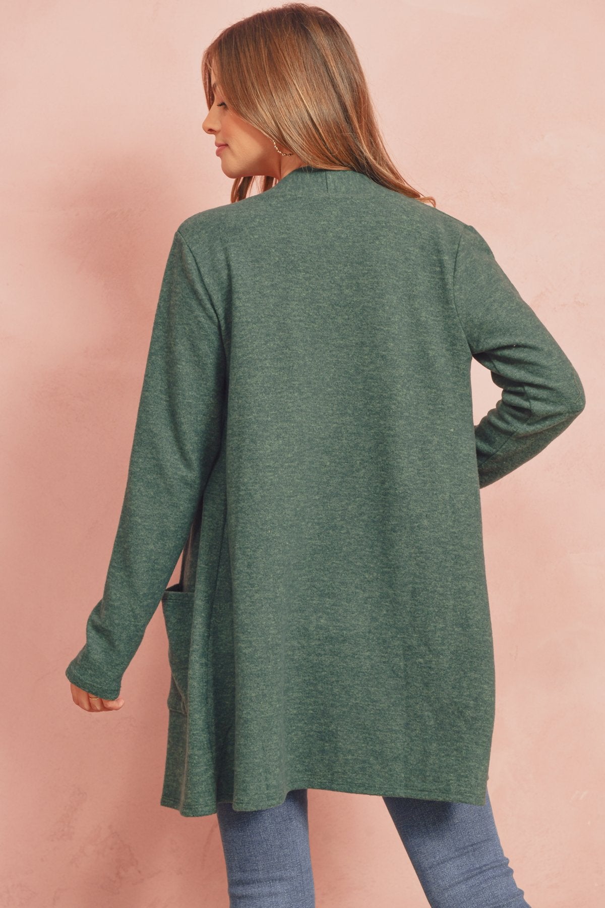 A woman in a Brushed Melange Open Front Cardigan by Riah Fashion and ripped jeans stands against a pink background, hands in her pockets.