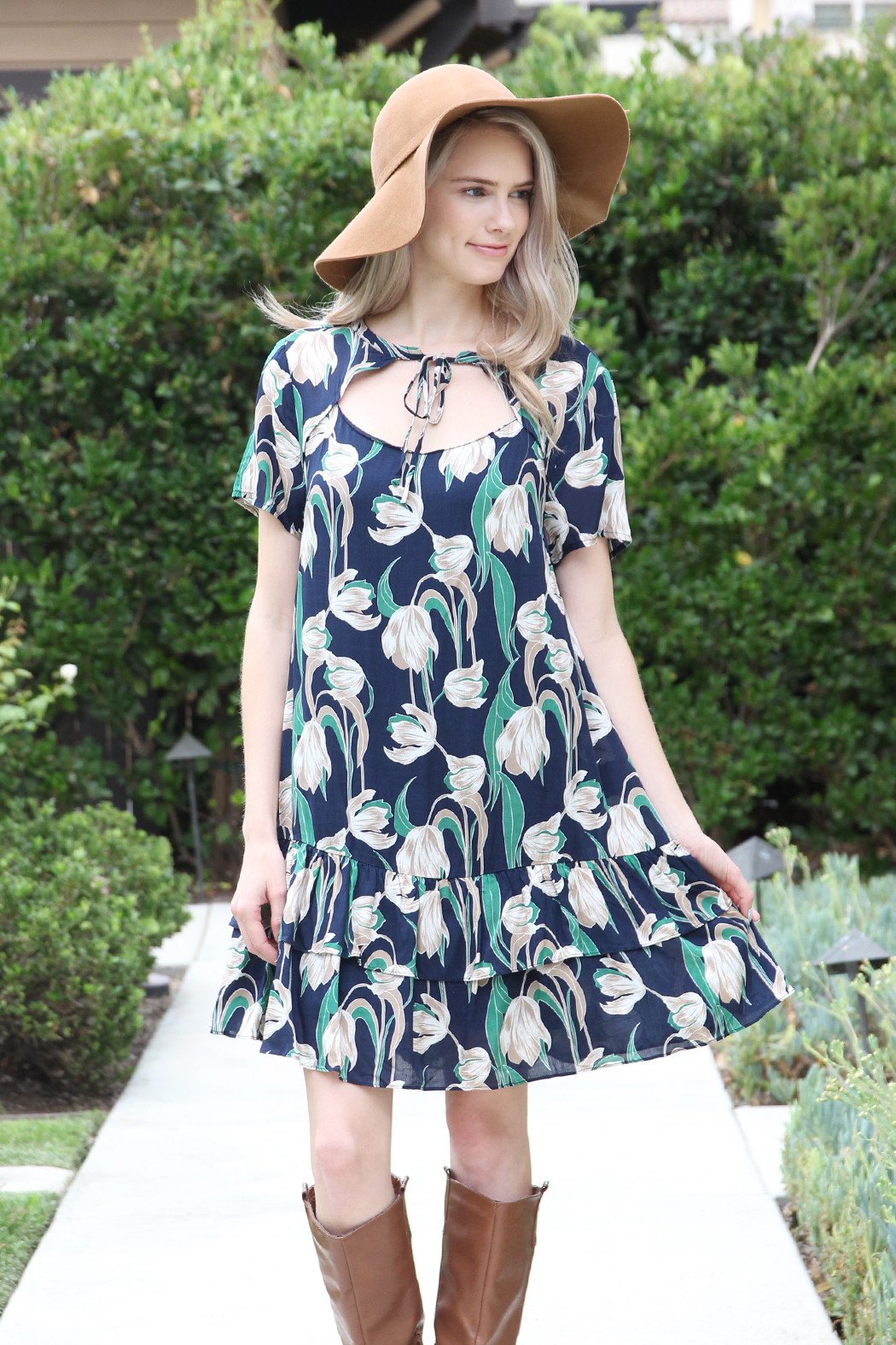A woman in a Riah Fashion Short Sleeve Floral Ruffle Dress and a brown hat stands near a wooden fence and greenery.