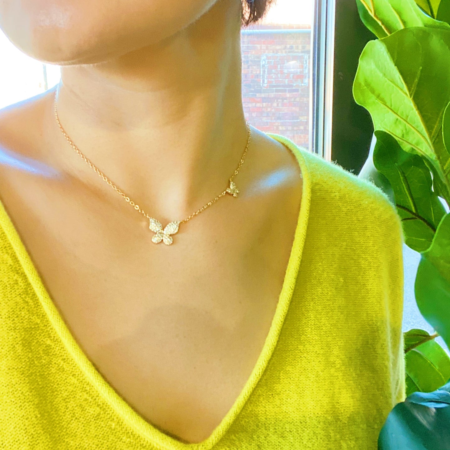 The Ellison + Young Double Shine Butterfly Necklace, featuring butterfly-shaped pendants adorned with small crystals, is elegantly displayed on a light marble background.