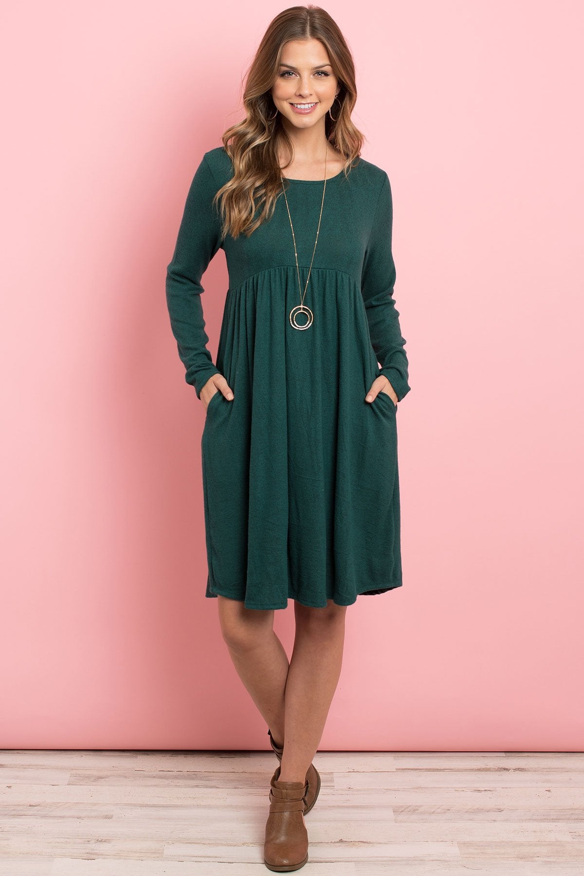 A woman in a Riah Fashion Boat Neck Brushed Hacci Round Hem Dress in Dark Ruby stands before a pink backdrop, adorned with a long necklace featuring circular pendants.