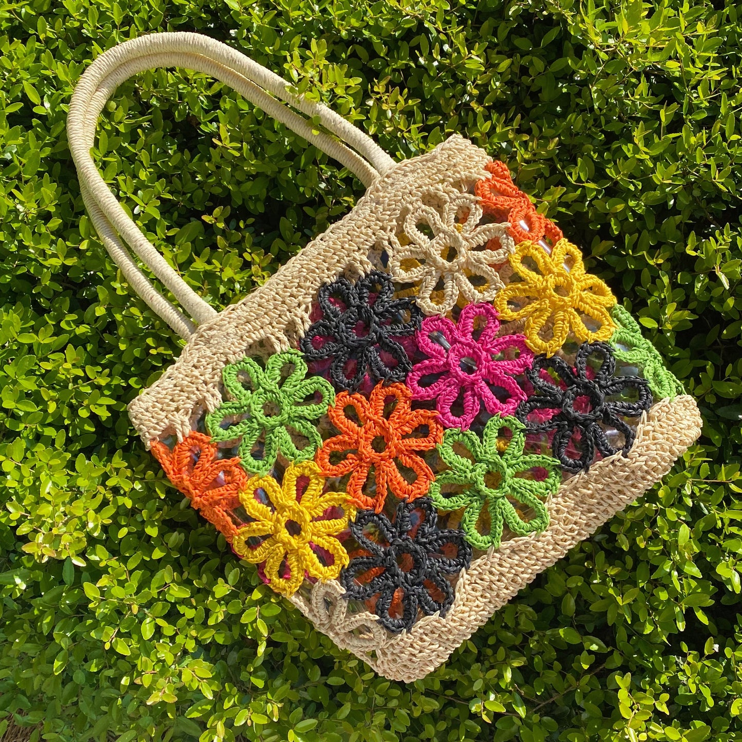The Hand Made Flower Power Tote by Ellison + Young, featuring colorful floral crochet patterns in orange, green, pink, black, and yellow, rests on dense green foliage.