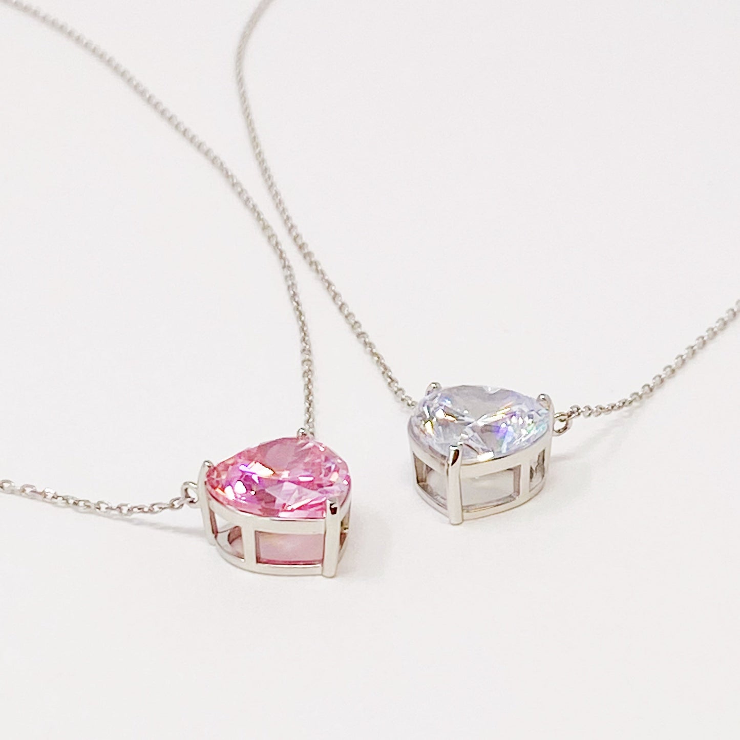 Two Absolute Beauty sterling silver heart necklaces by Ellison + Young; one with a pink gem, the other with a clear gem, against a white background.