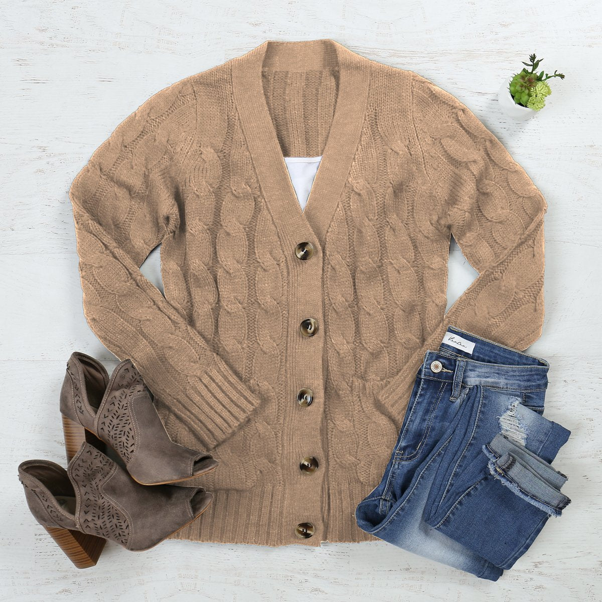 A person wearing an ash copper cable sweater with pockets from Riah Fashion stands in front of tall green plants and a wooden fence.