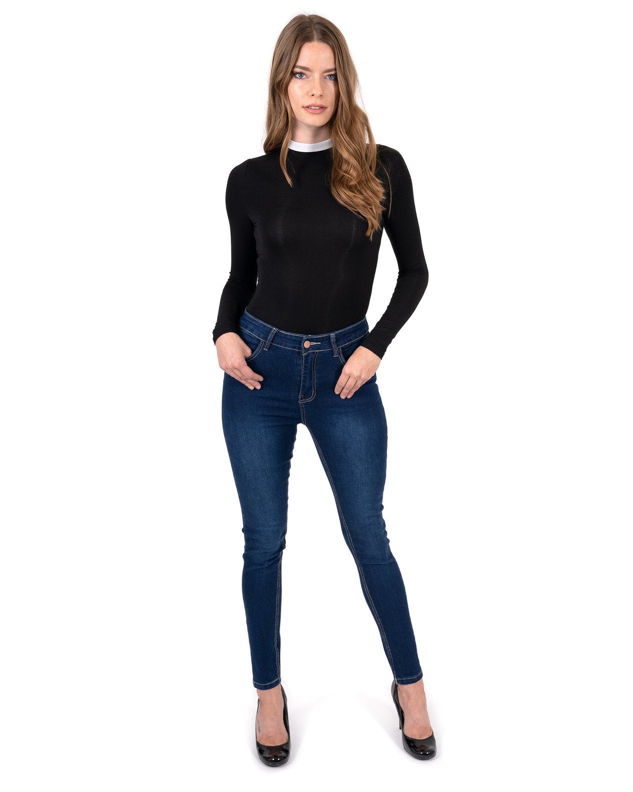 A person stands by a pool wearing a black long-sleeve shirt, Alexis High Waist Skinny Jeans by Savoy Active, and heels. Tall trees and vibrant flowers surround the area.