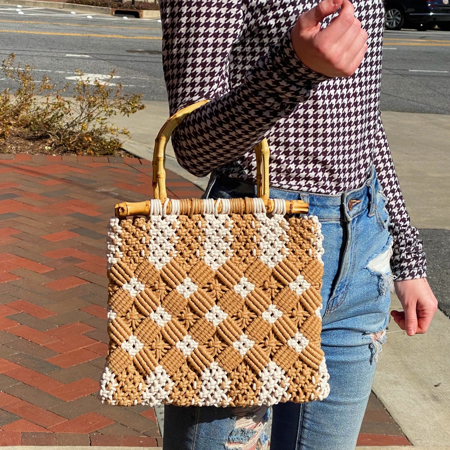A person holds the Ellison + Young Bamboo Babe Tote, wearing a houndstooth shirt and ripped jeans. The background features a paved walkway and street.