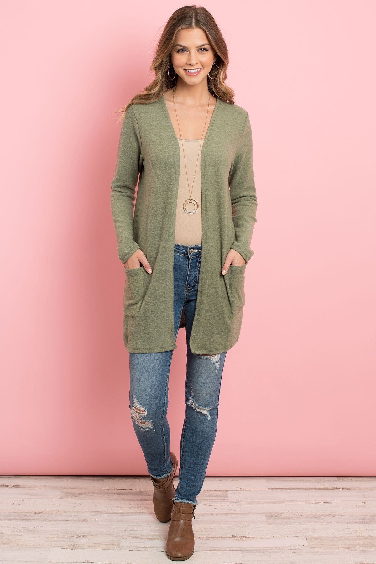 A woman in a teal Solid Long Sleeved Open Front Pocket Cardigan by Riah Fashion over a beige top stands against a pink background, smiling with hands in pockets.