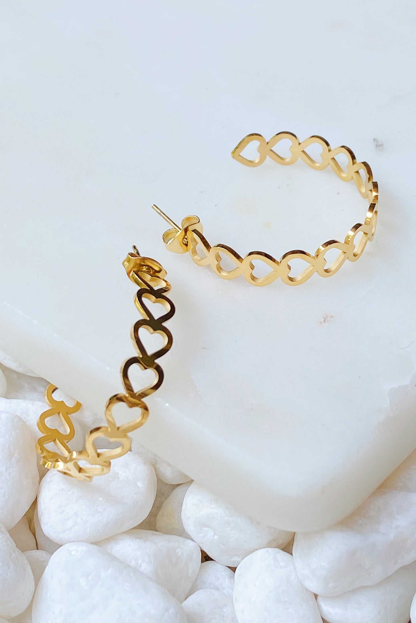 Ellison + Youngs Linked Heart Hoop Earrings, featuring heart-shaped cutouts, are elegantly displayed on a white marble surface surrounded by white pebbles.
