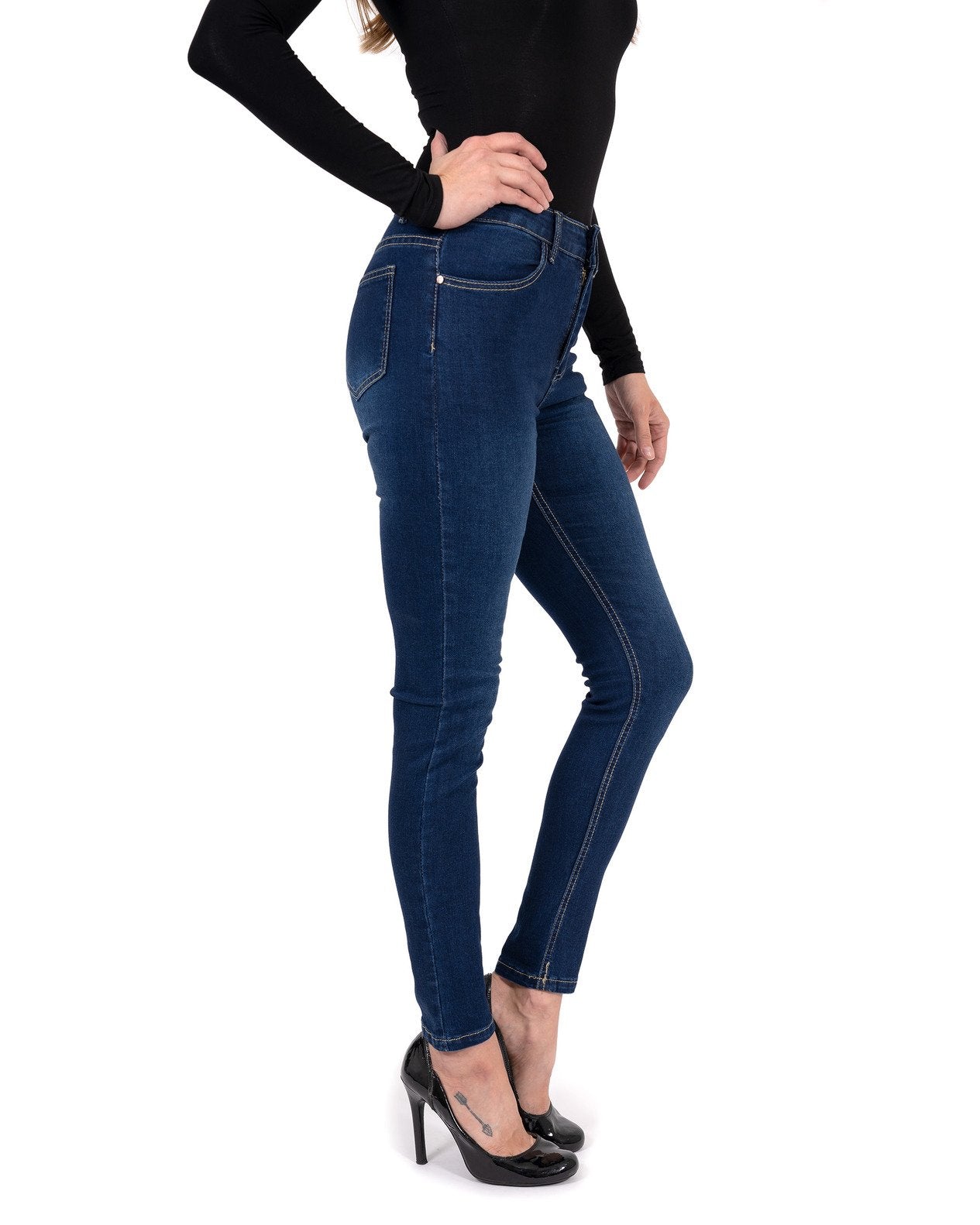 A person stands by a pool wearing a black long-sleeve shirt, Alexis High Waist Skinny Jeans by Savoy Active, and heels. Tall trees and vibrant flowers surround the area.