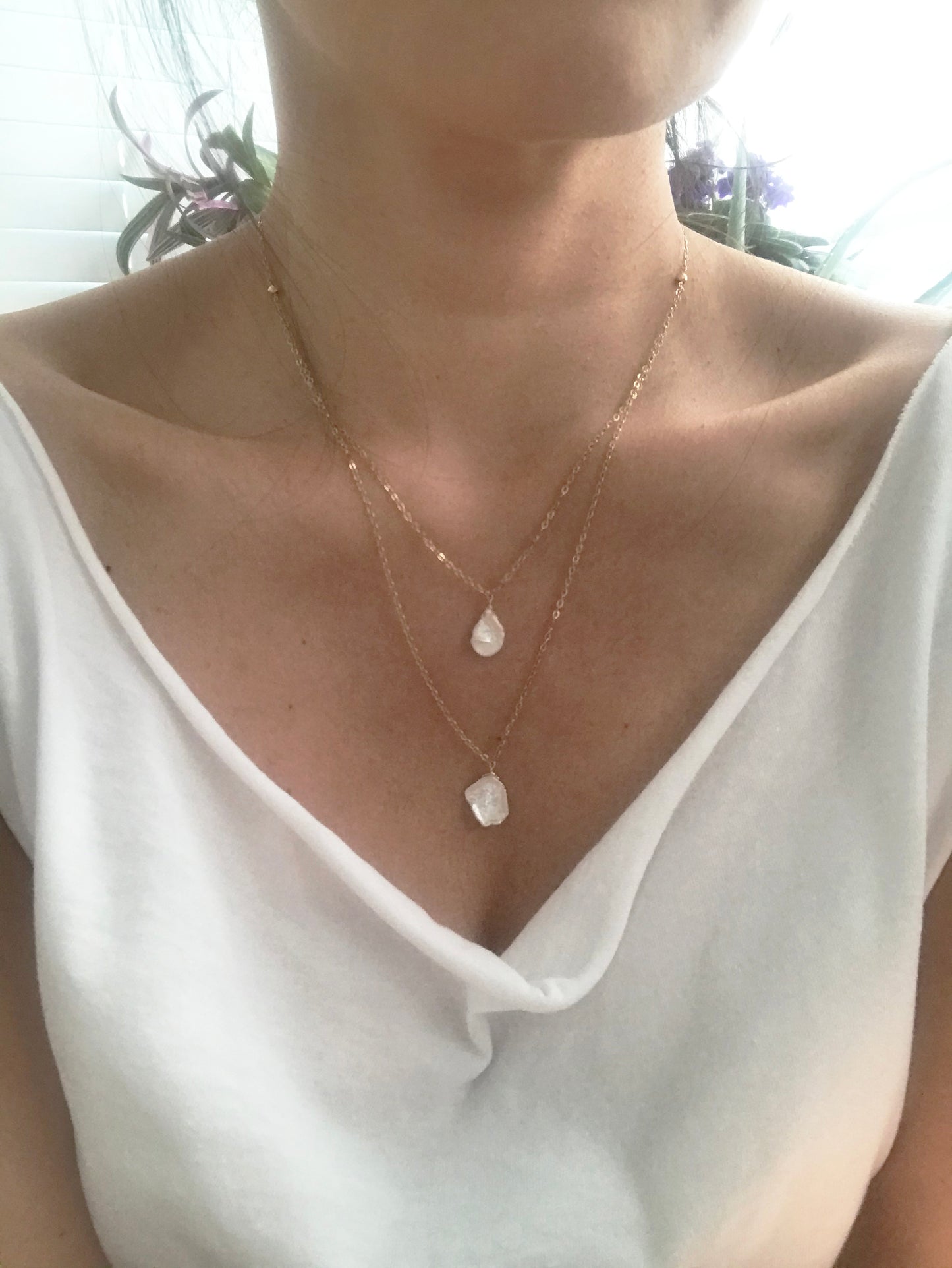 The Keshi Pearl Layered Necklace by Gena Myint is a gold double-layer necklace featuring two clear gemstone pendants: one pear-shaped and the other rectangular.
