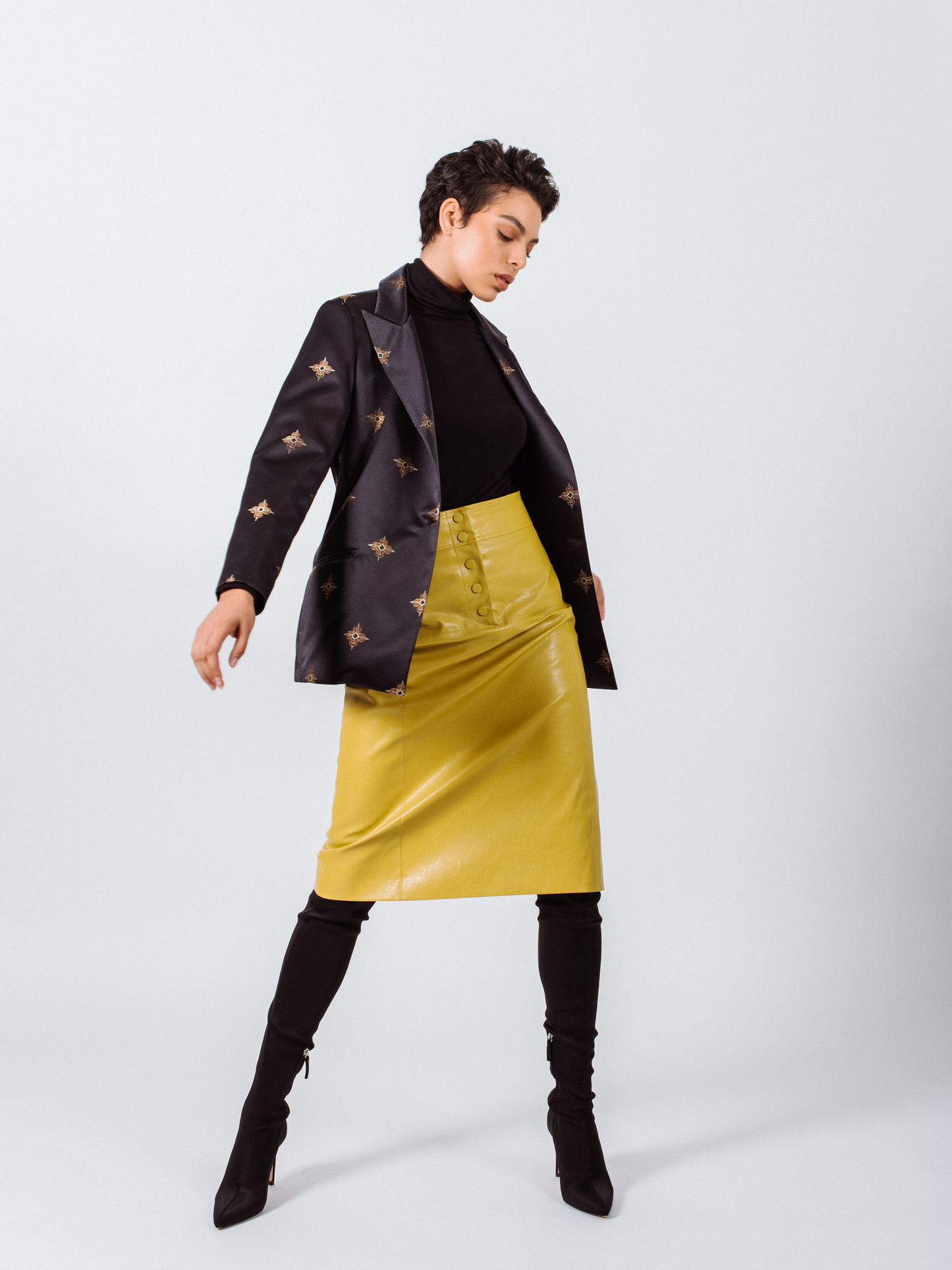 Person posing in a short-sleeve black shirt and black ankle boots, wearing Le Réussi®s Power Woman- Mustard Leather Skirt, in front of a plain white background.