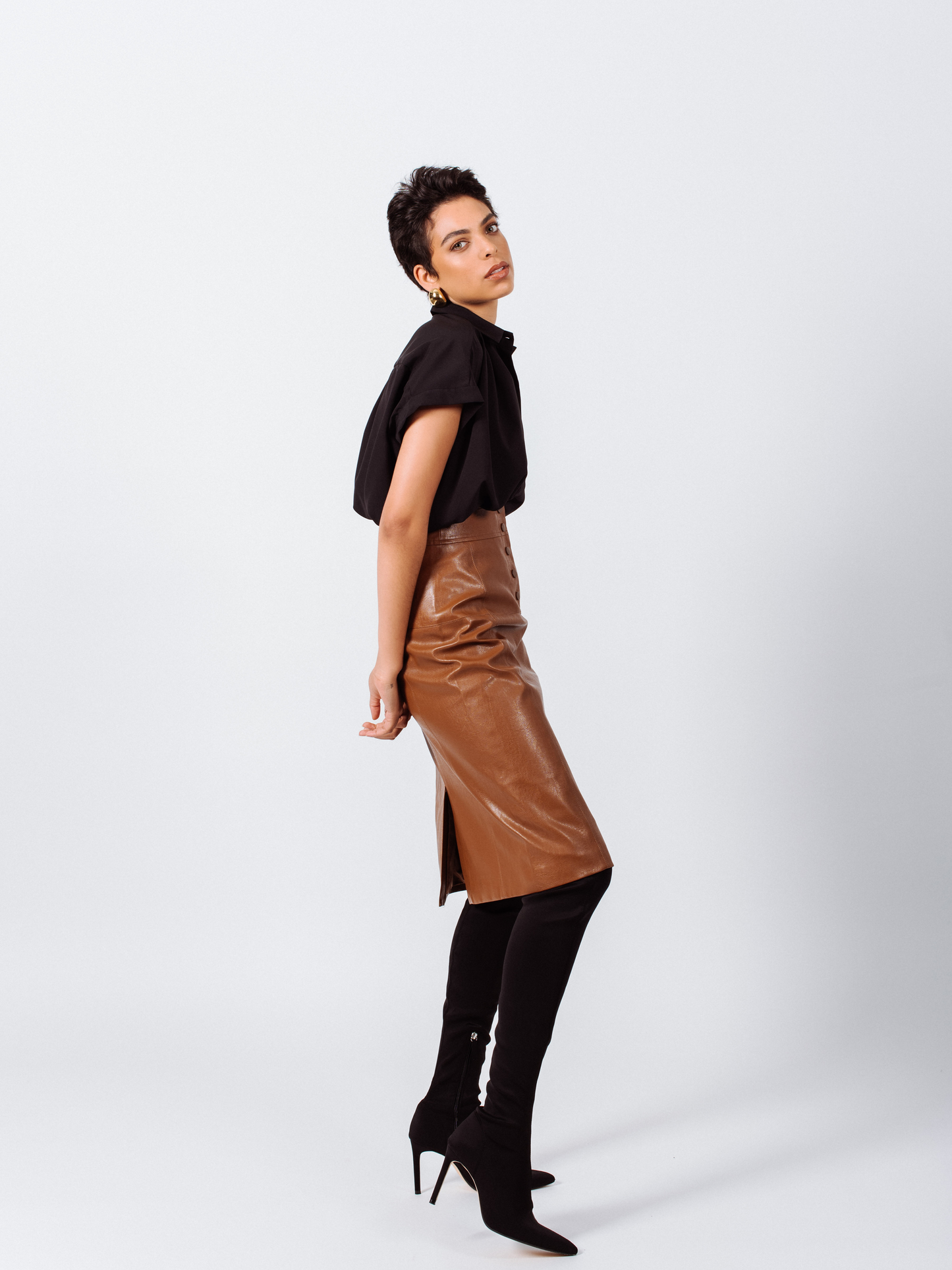 A woman in a black blouse, Power Woman- Dark Hazel Leather Skirt by Le Réussi®, and black ankle boots stands against a plain white background.
