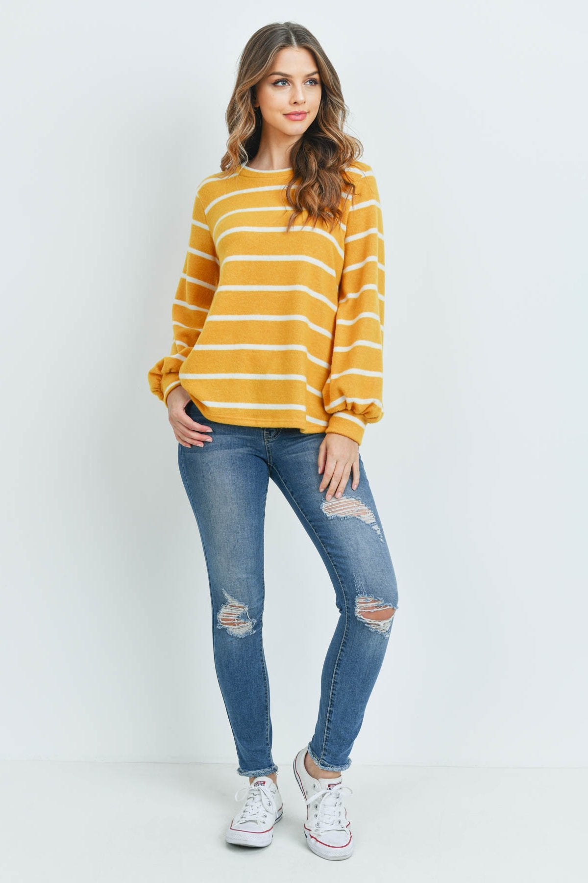 A woman in a Riah Fashion Striped Long Sleeved Round Neck Swing Top and jeans stands against a pink backdrop, wearing a long necklace and touching her hair.