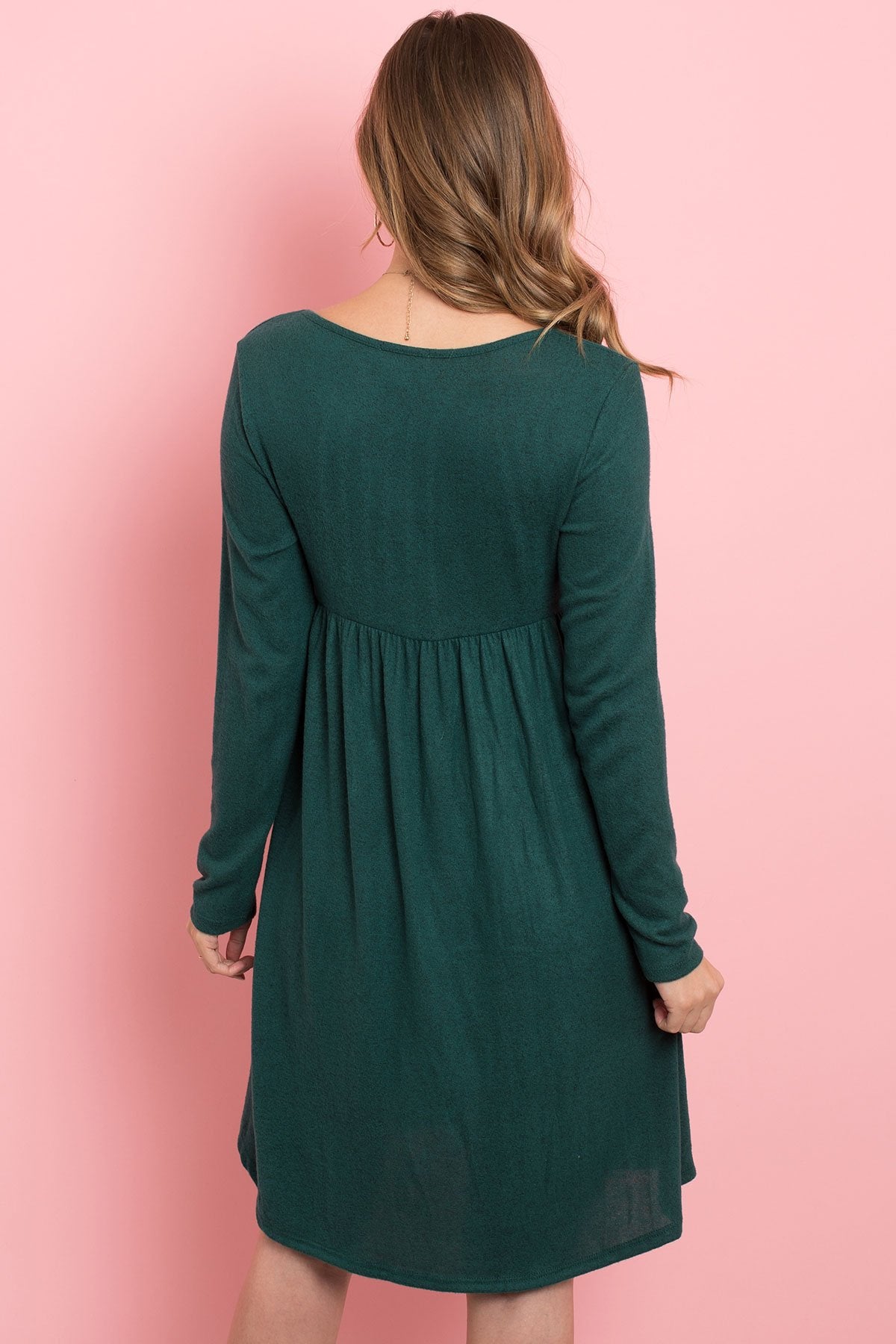 A woman in a Riah Fashion Boat Neck Brushed Hacci Round Hem Dress in Dark Ruby stands before a pink backdrop, adorned with a long necklace featuring circular pendants.