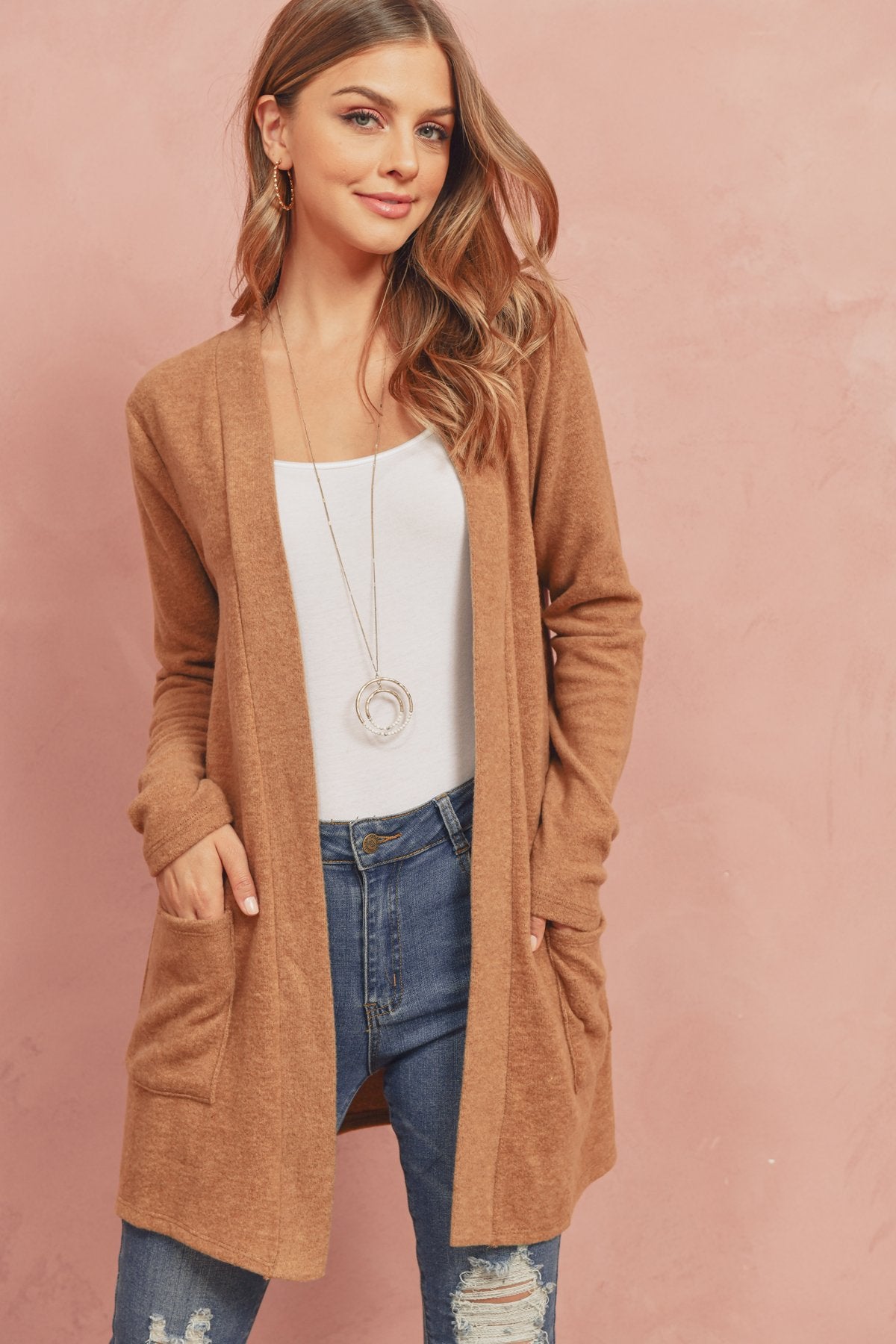 A woman in a Brushed Melange Open Front Cardigan by Riah Fashion and ripped jeans stands against a pink background, hands in her pockets.