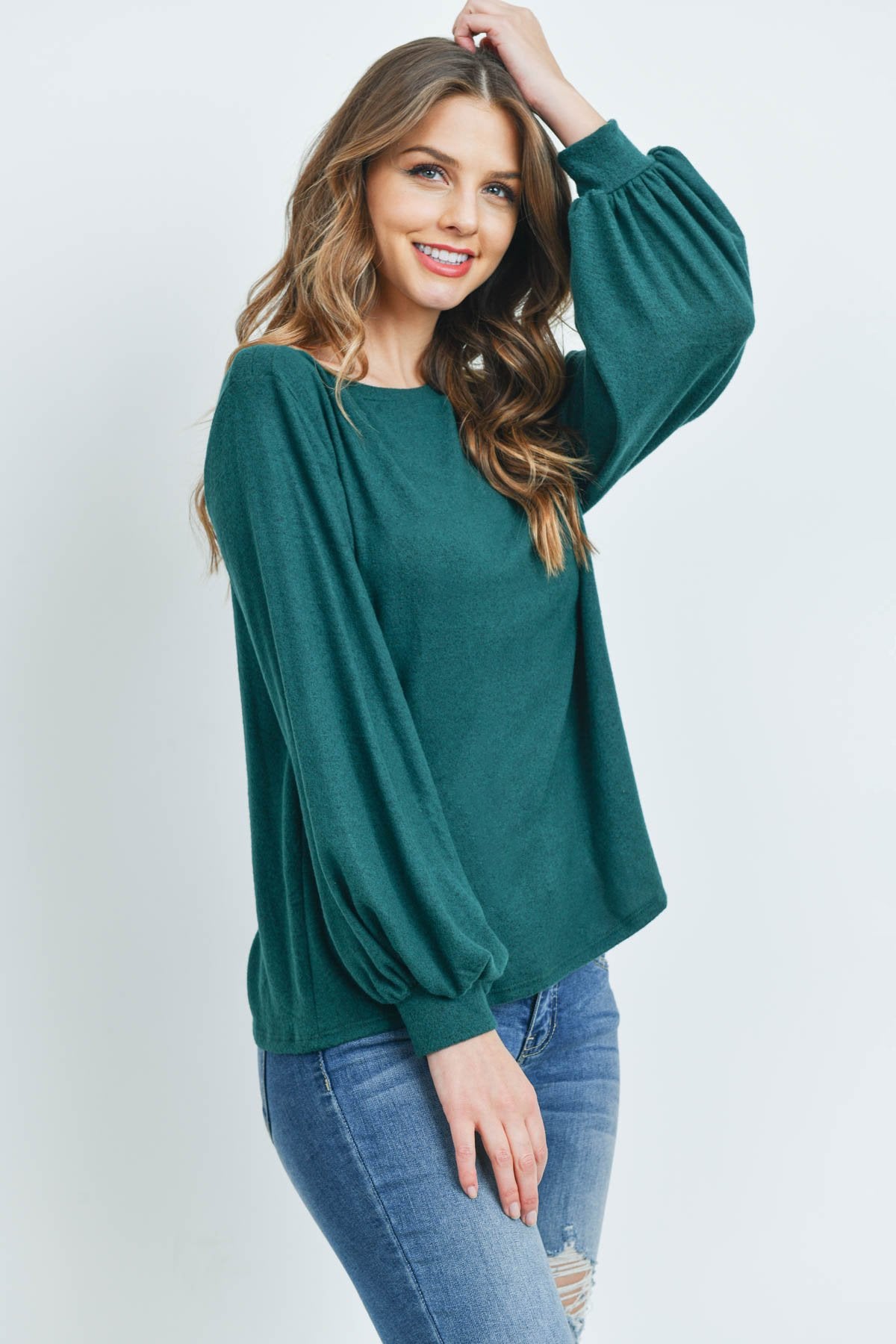 A woman models a Riah Fashion Puff Sleeved Boat Neck Two Toned Brushed Hacci Top with ripped jeans and brown boots on a concrete floor against a plain backdrop, accessorized with a long necklace.