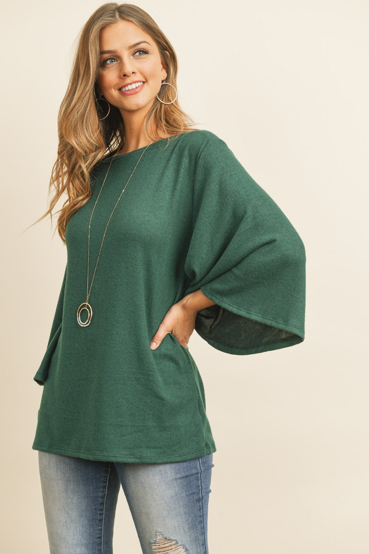 A person in a Riah Fashion Boat Neck Bell Sleeve Solid Hacci Brushed Top and ripped jeans stands against a neutral background, accessorized with hoop earrings and a long necklace.