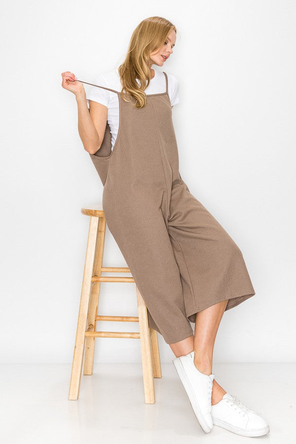 A person with long blonde hair sits on a wooden stool against a plain background, wearing Stylespects Cropped Bottom Wide Leg Oversized Jumpsuit in taupe and white sneakers.