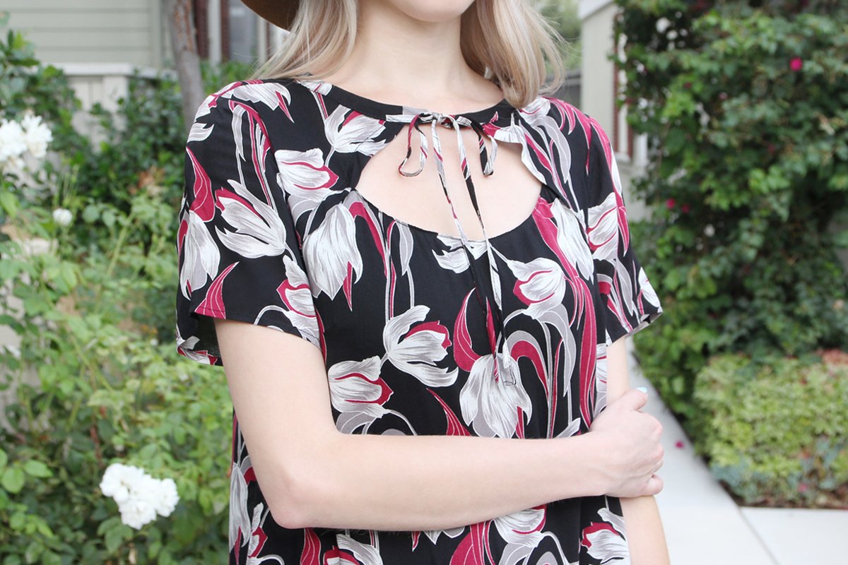 A woman in a Riah Fashion Short Sleeve Floral Ruffle Dress and a brown hat stands near a wooden fence and greenery.