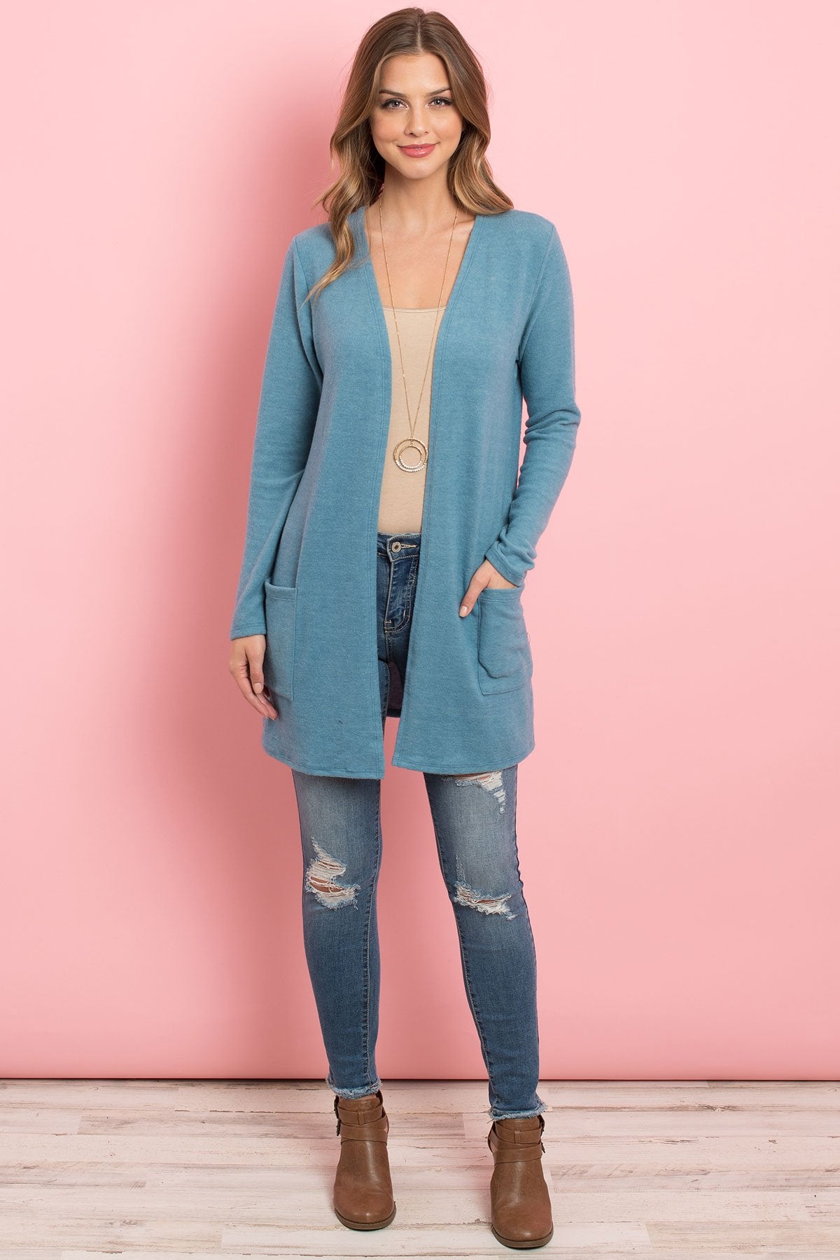 A woman in a teal Solid Long Sleeved Open Front Pocket Cardigan by Riah Fashion over a beige top stands against a pink background, smiling with hands in pockets.