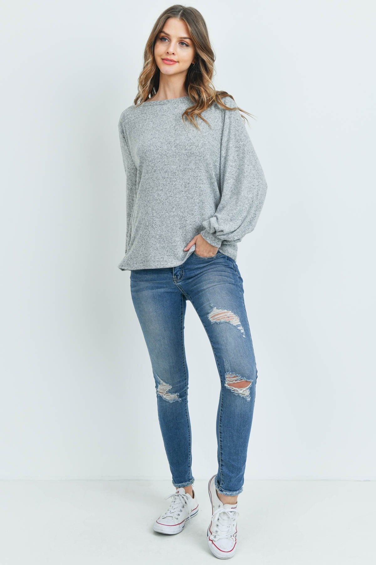 A woman models a Riah Fashion Puff Sleeved Boat Neck Two Toned Brushed Hacci Top with ripped jeans and brown boots on a concrete floor against a plain backdrop, accessorized with a long necklace.