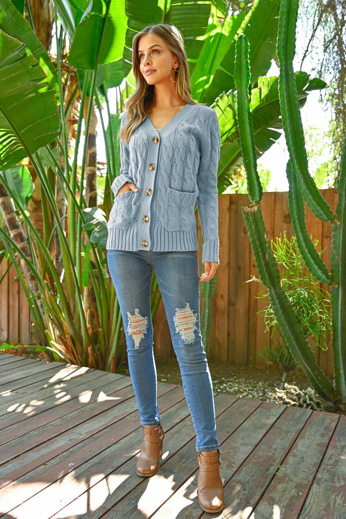 A person wearing an ash copper cable sweater with pockets from Riah Fashion stands in front of tall green plants and a wooden fence.