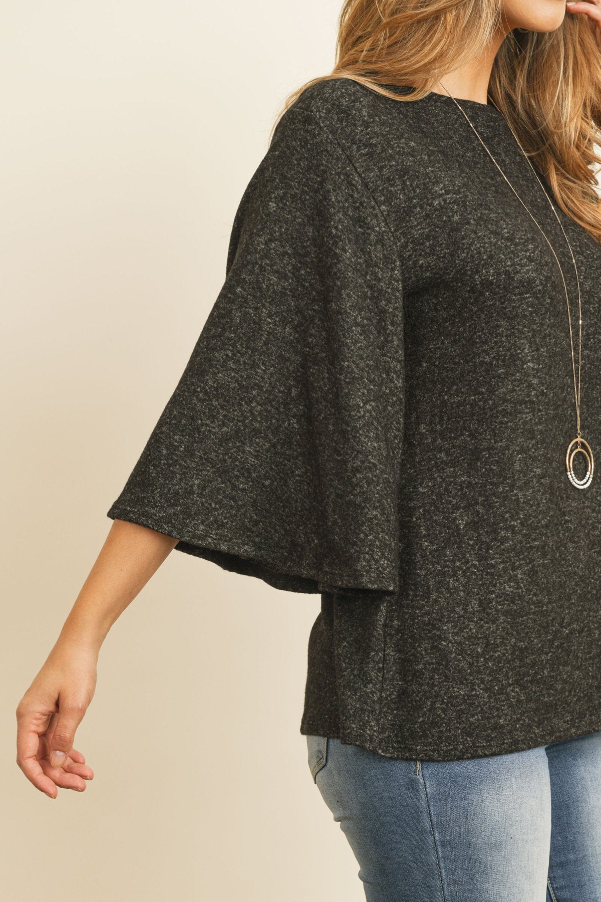 A person in a Riah Fashion Boat Neck Bell Sleeve Solid Hacci Brushed Top and ripped jeans stands against a neutral background, accessorized with hoop earrings and a long necklace.
