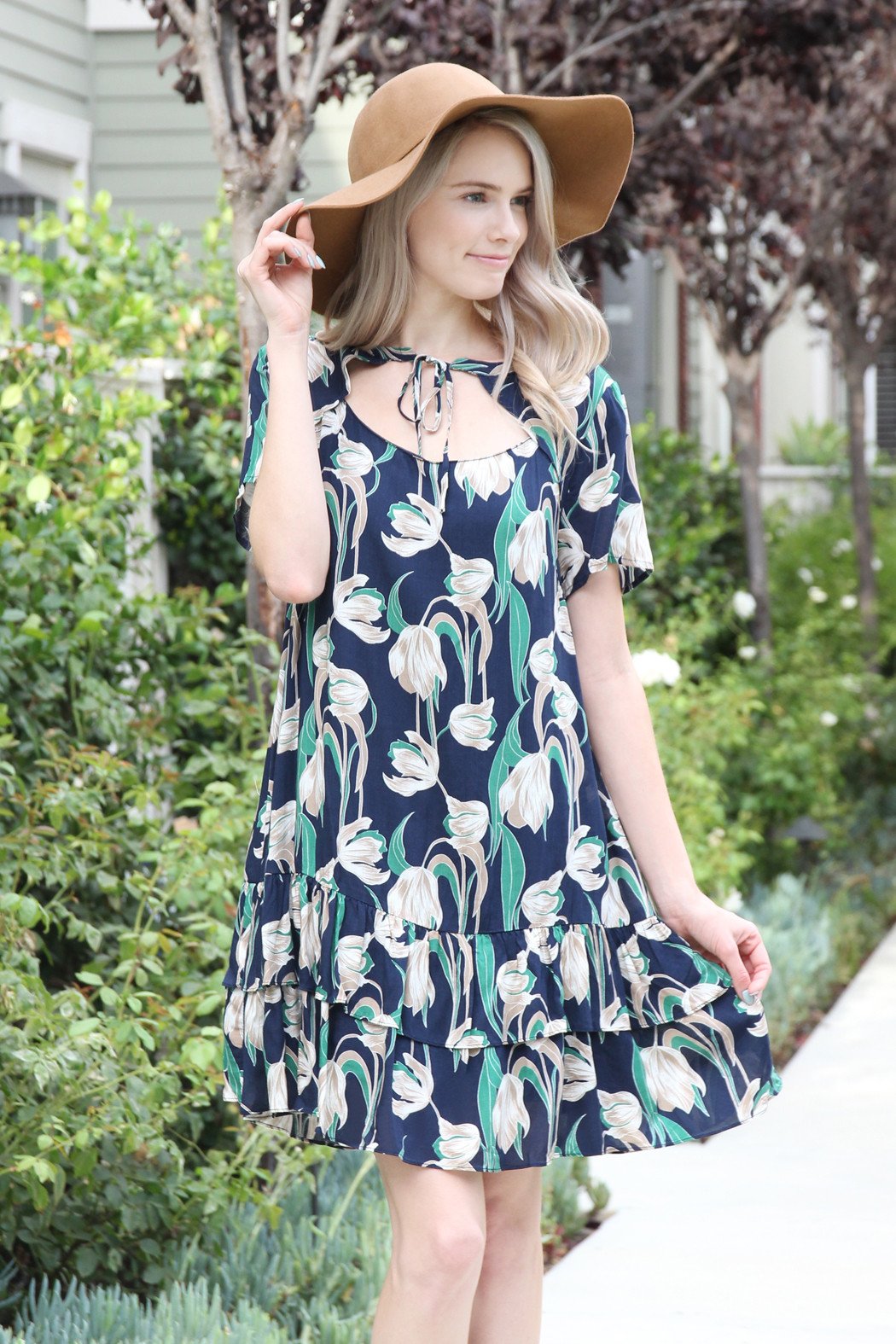 A woman in a Riah Fashion Short Sleeve Floral Ruffle Dress and a brown hat stands near a wooden fence and greenery.