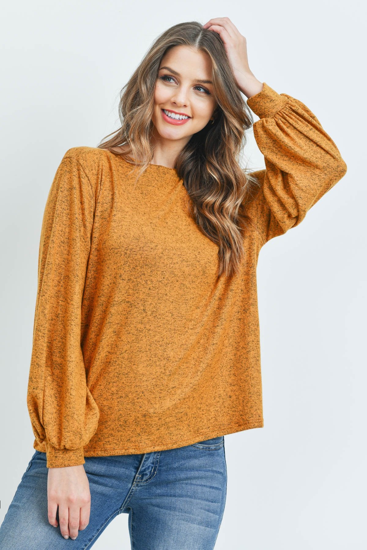 A woman models a Riah Fashion Puff Sleeved Boat Neck Two Toned Brushed Hacci Top with ripped jeans and brown boots on a concrete floor against a plain backdrop, accessorized with a long necklace.